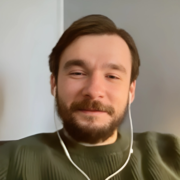 A man with light skin, dark brown hair, and a well-groomed beard is seen smiling gently while wearing white earphones. He is dressed in a dark green sweater with a textured pattern, sitting in a relaxed posture against a neutral-toned background. The image has a warm, soft focus.
