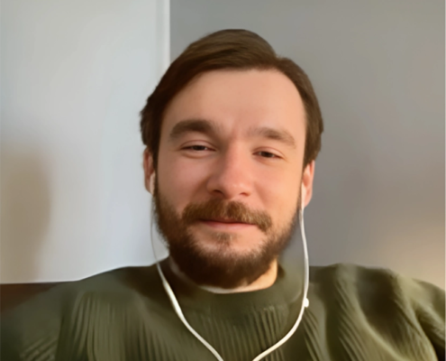 A man with light skin, dark brown hair, and a well-groomed beard is seen smiling gently while wearing white earphones. He is dressed in a dark green sweater with a textured pattern, sitting in a relaxed posture against a neutral-toned background. The image has a warm, soft focus.