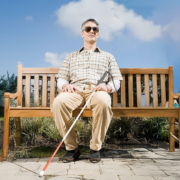 A blind man with short gray hair sits on a wooden bench in a park under a bright blue sky with scattered clouds. He wears sunglasses, a plaid short-sleeved shirt over a white long-sleeve shirt, beige pants, and black shoes. He holds a white cane with a red tip in his hands, resting it on the ground in front of him, while maintaining a relaxed yet upright posture.