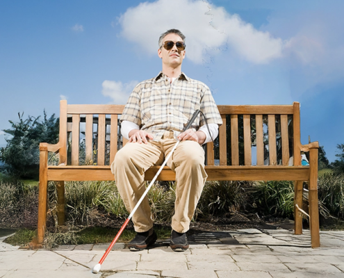 A blind man with short gray hair sits on a wooden bench in a park under a bright blue sky with scattered clouds. He wears sunglasses, a plaid short-sleeved shirt over a white long-sleeve shirt, beige pants, and black shoes. He holds a white cane with a red tip in his hands, resting it on the ground in front of him, while maintaining a relaxed yet upright posture.